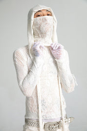 A person looks directly at the camera, wearing a semi-sheer white lace top with a matching lace hood mask and lace gloves