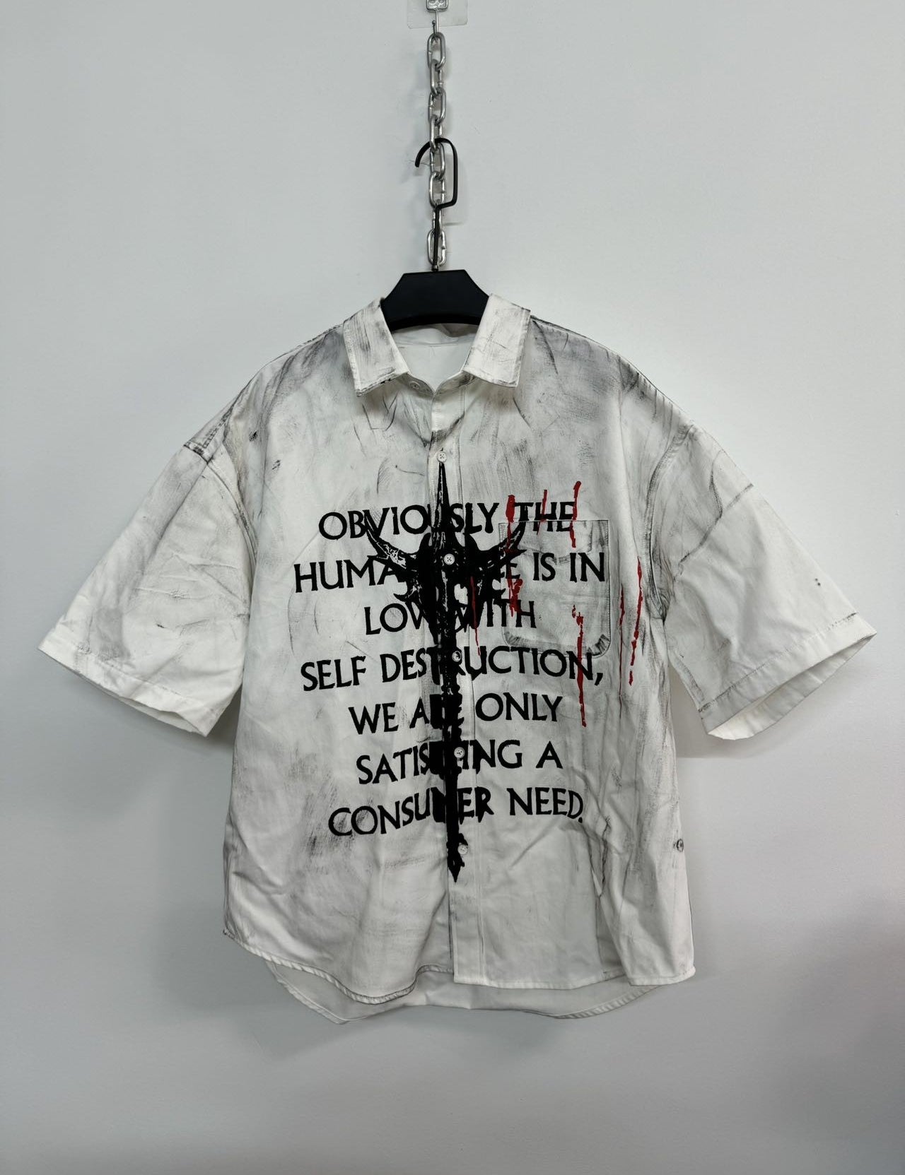 A white men's graffiti t-shirt with short sleeves, featuring black text and a central abstract black graphic, along with red paint splatters, hanging on a white wall.