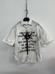 A white men's graffiti t-shirt with short sleeves, featuring black text and a central abstract black graphic, along with red paint splatters, hanging on a white wall.