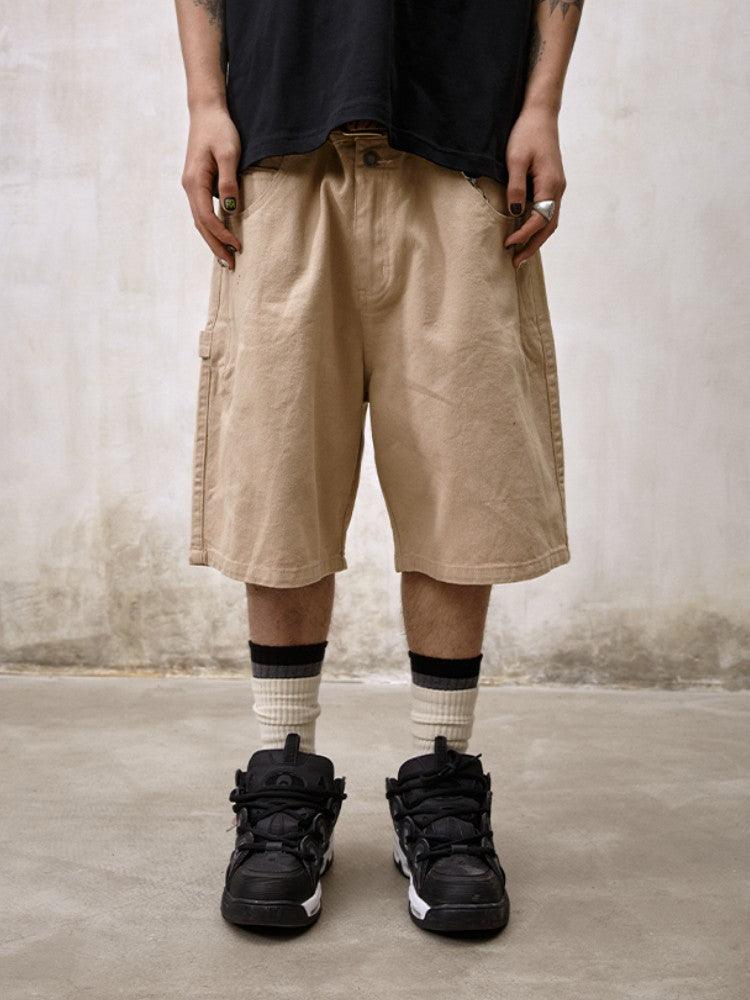 A person stands facing the camera, wearing Remedy Straight Workwear Mid-Length Jorts in a khaki color, paired with a black T-shirt, striped white socks, and black and white sneakers.