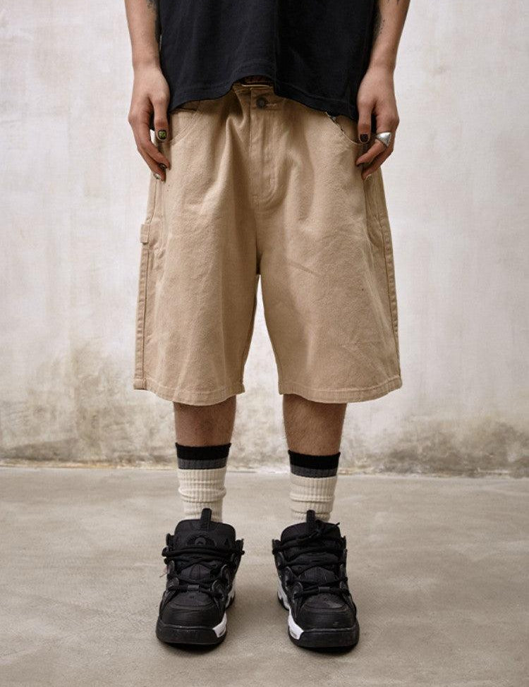 A person stands facing the camera, wearing Remedy Straight Workwear Mid-Length Jorts in a khaki color, paired with a black T-shirt, striped white socks, and black and white sneakers.