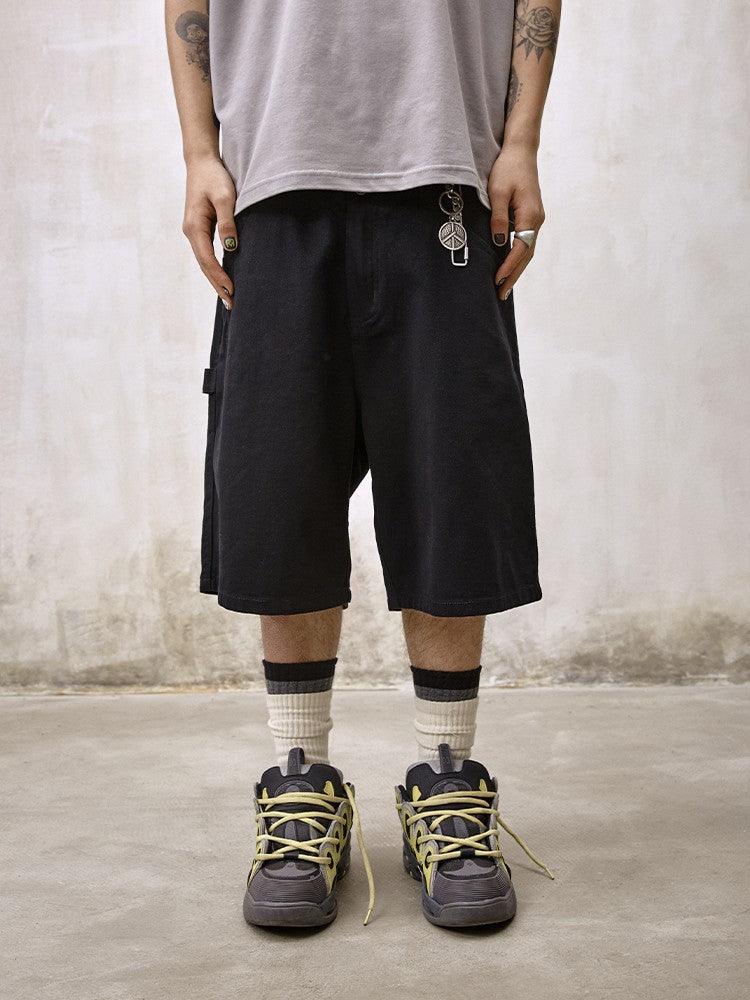 A person stands facing forward, wearing Remedy Straight Workwear Mid-Length Jorts in black, a light gray t-shirt, and chunky grey sneakers with vibrant yellow laces and white and black striped socks, accessorized with a peace sign keychain.