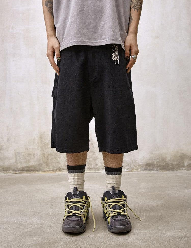 A person stands facing forward, wearing Remedy Straight Workwear Mid-Length Jorts in black, a light gray t-shirt, and chunky grey sneakers with vibrant yellow laces and white and black striped socks, accessorized with a peace sign keychain.