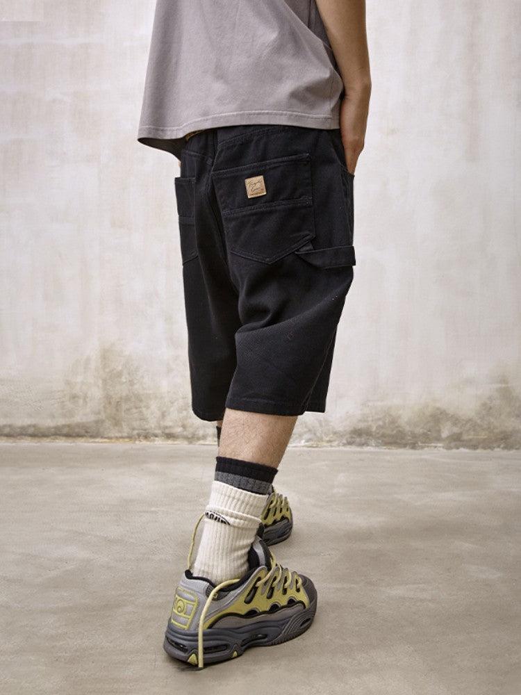 A black pair of Remedy Straight Workwear Mid-Length Jorts is shown on a mannequin and modeled by a person from behind, who is also wearing a gray t-shirt and chunky gray and yellow sneakers with striped socks.