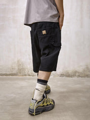 A black pair of Remedy Straight Workwear Mid-Length Jorts is shown on a mannequin and modeled by a person from behind, who is also wearing a gray t-shirt and chunky gray and yellow sneakers with striped socks.
