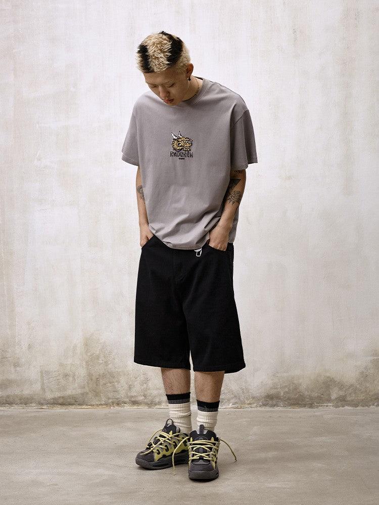 A person with dyed hair and tattoos stands with their hands in their pockets, wearing a gray graphic t-shirt, Remedy Straight Workwear Mid-Length Jorts in black, and chunky gray and yellow sneakers with striped socks.