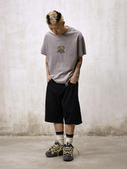 A person with dyed hair and tattoos stands with their hands in their pockets, wearing a gray graphic t-shirt, Remedy Straight Workwear Mid-Length Jorts in black, and chunky gray and yellow sneakers with striped socks.