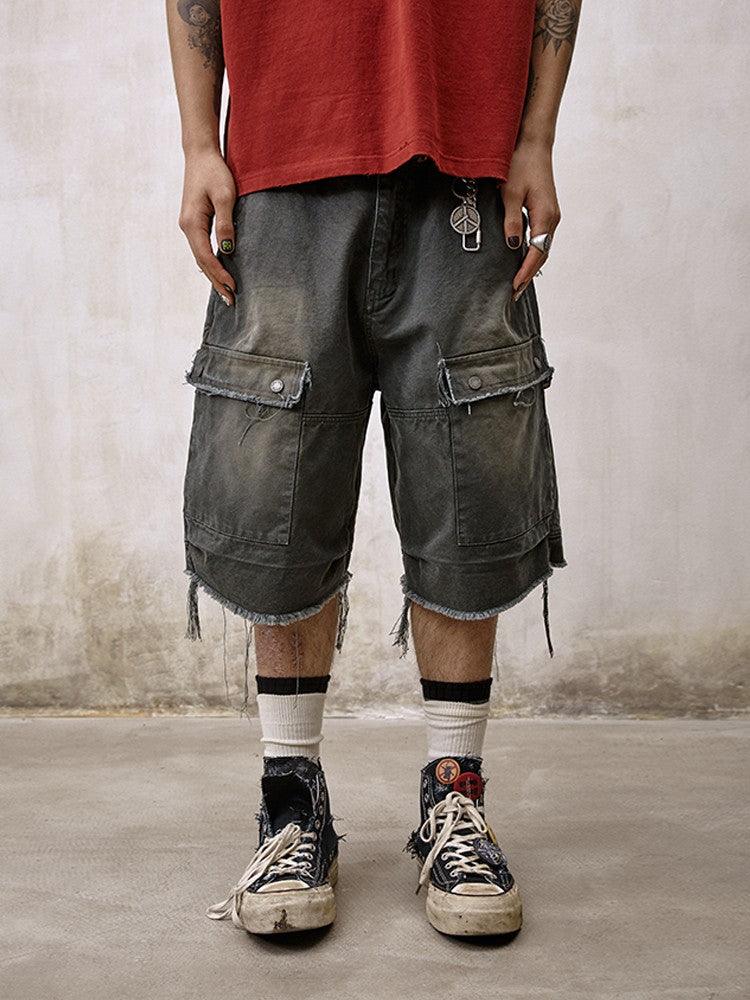 A person stands wearing distressed and frayed Remedy Washed Tassel Workwear Jorts with large cargo pockets, paired with a faded red T-shirt and dirty sneakers with white socks.