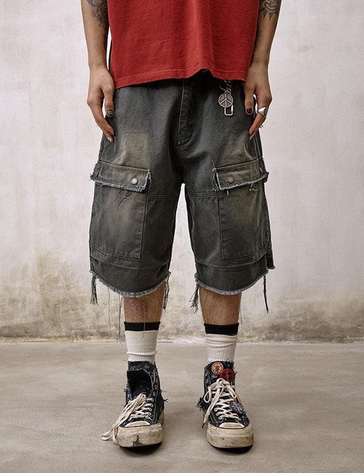 A person stands wearing distressed and frayed Remedy Washed Tassel Workwear Jorts with large cargo pockets, paired with a faded red T-shirt and dirty sneakers with white socks.