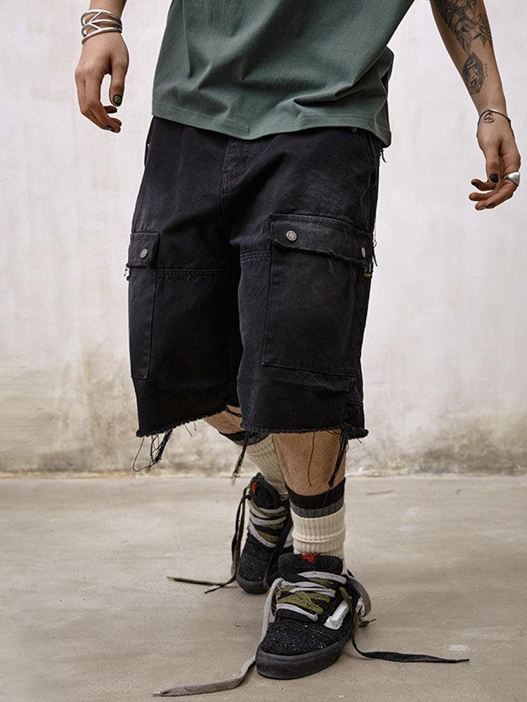 A person stands wearing distressed black Remedy Washed Tassel Workwear Jorts with frayed hems and cargo pockets, a green t-shirt, and dirty black sneakers with white and black striped socks.