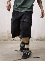 A person stands wearing distressed black Remedy Washed Tassel Workwear Jorts with frayed hems and cargo pockets, a green t-shirt, and dirty black sneakers with white and black striped socks.