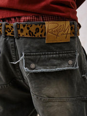 A close-up shot of the back of Remedy Washed Tassel Workwear Jorts reveals a distinctive leopard print belt, a brown leather "Remedy" brand patch, and a distressed back pocket with metal buttons.
