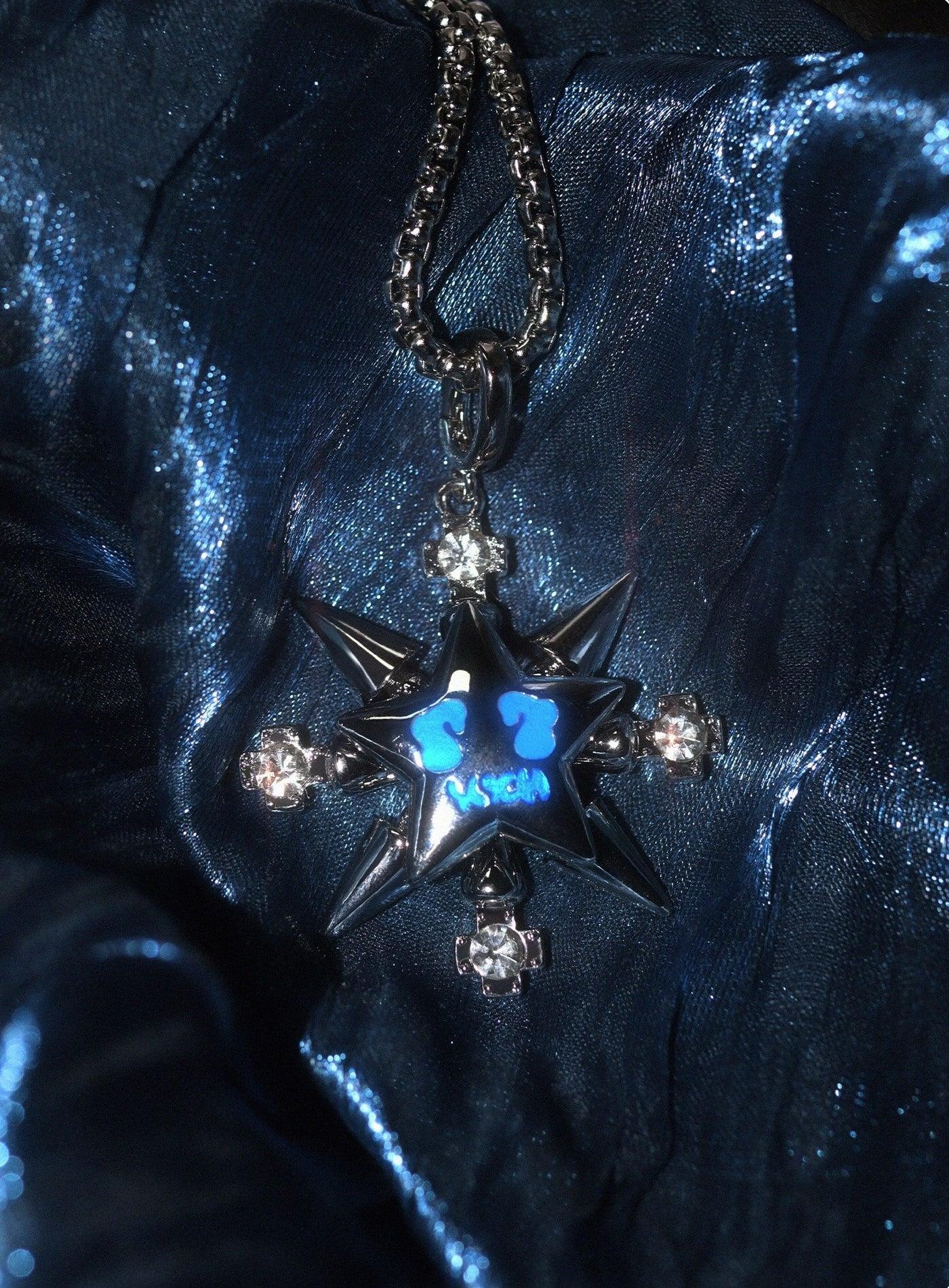 A shiny silver "VSCH Lucky Star Necklace" with a star pendant and blue and diamond accents rests on a reflective blue surface.