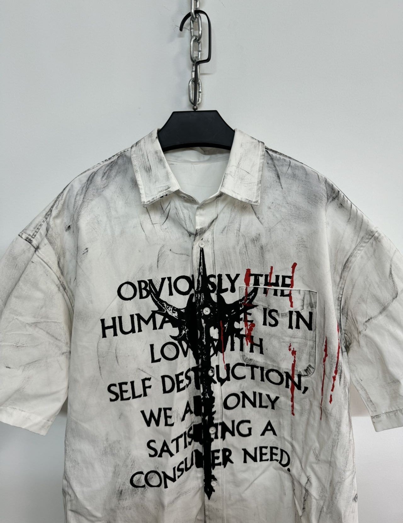 A close-up of a white men's graffiti t-shirt with short sleeves, featuring black text, a central abstract black graphic, and red paint splatters, hanging on a white wall.





