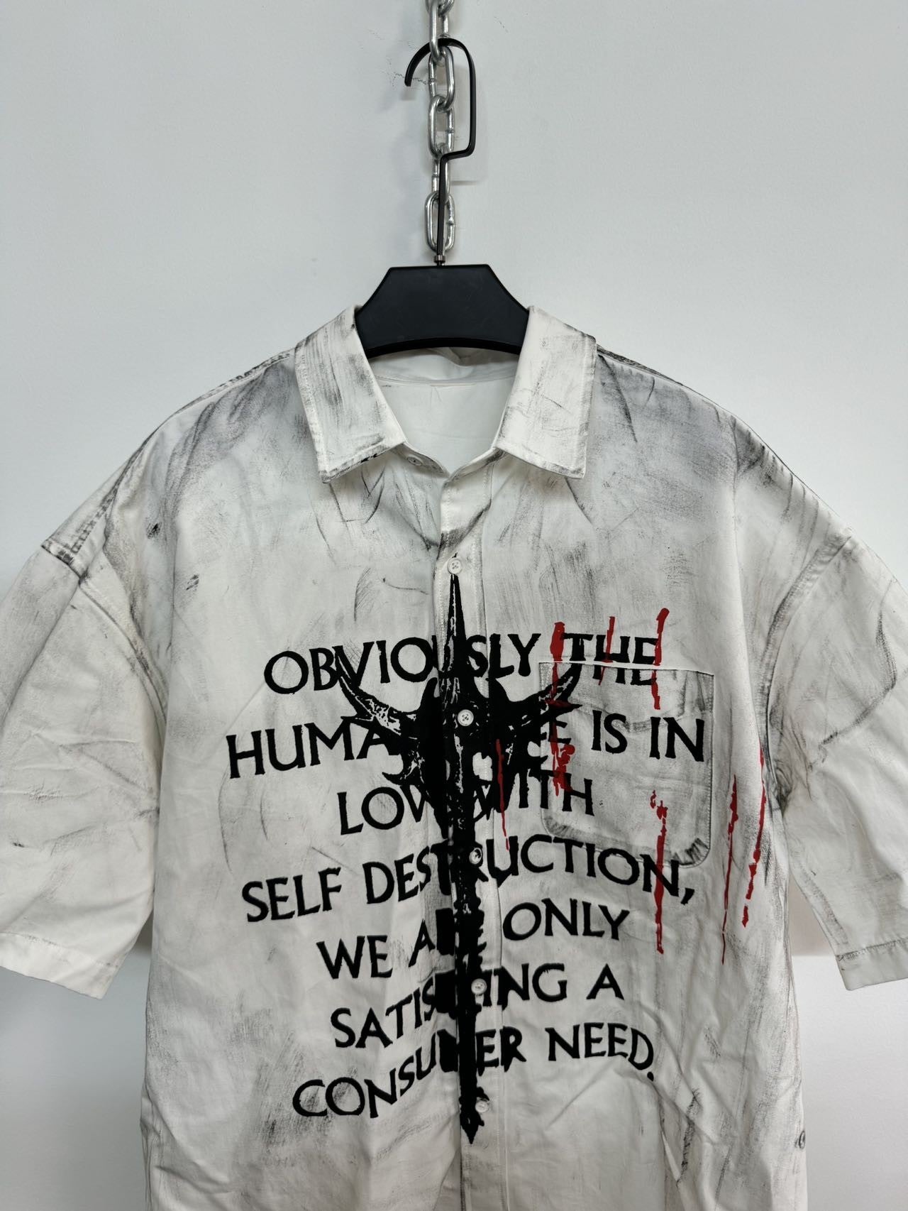 A close-up of a white men's graffiti t-shirt with short sleeves, featuring black text, a central abstract black graphic, and red paint splatters, hanging on a white wall.





