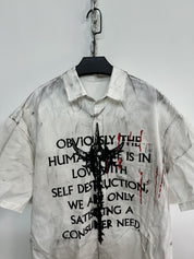 A close-up of a white men's graffiti t-shirt with short sleeves, featuring black text, a central abstract black graphic, and red paint splatters, hanging on a white wall.





