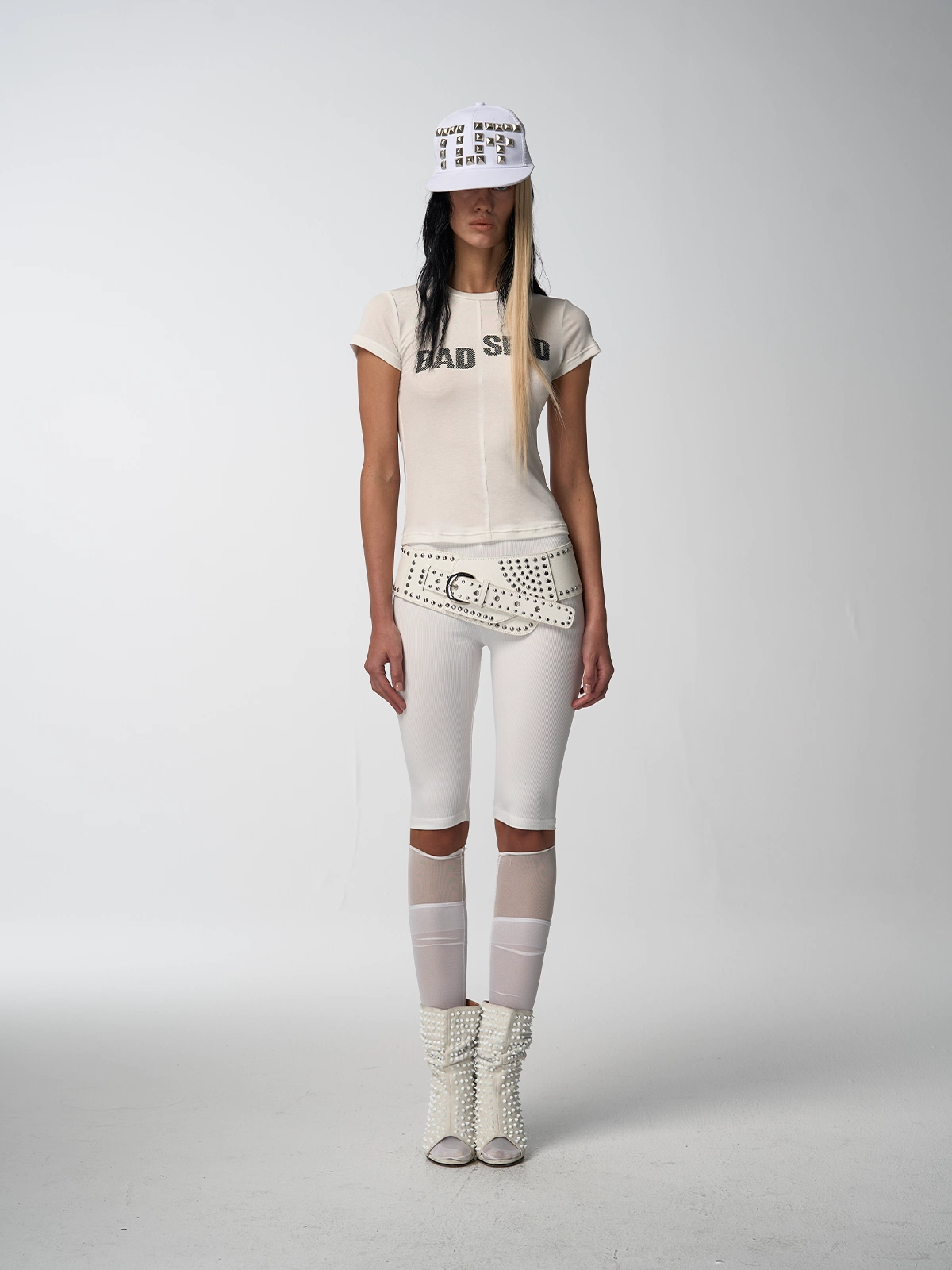 A full-length shot of a woman with Women's Rebel tee shirt," white studded belt, white ribbed pants with sheer knee details.