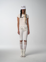 A full-length shot of a woman with Women's Rebel tee shirt," white studded belt, white ribbed pants with sheer knee details.